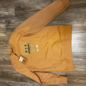 Beige sweatshirt från Burberry M - Säljer en snygg beige sweatshirt från Burberry i storlek M. Tröjan har en stor patch med Burberry-logga och tryck på bröstet samt ribbade muddar vid ärmslut och nederkant. Perfekt för dig som gillar stilrena och exklusiva plagg med streetkänsla.