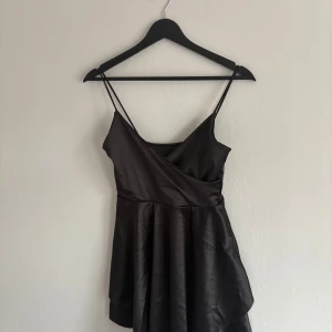 Svart omlottklänning i satin - Supersnygg svart kortklänning i satin med omlottliv och tunna axelband. Klänningen har en lätt glansig finish och en flowy kjol som ger en elegant vibe. Perfekt för dig som gillar minimalistisk stil med en touch av lyx.