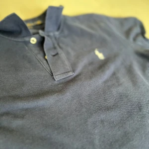 Marinblå pikétröja från Ralph Lauren - Klassisk marinblå pikétröja från Ralph Lauren med vit broderad logga på bröstet. Tröjan har korta ärmar, krage och knäppning med två knappar. Tillverkad i bomull för en skön och luftig känsla. Perfekt för en stilren och avslappnad look. Storlek s men passar ungefär som xs
