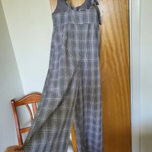 Rutig byxdress från Reclaimed Vintage - Snygg rutig byxdress från Reclaimed Vintage i vitt,grått och lila med breda axelband som knyts och dragkedja i ryggen. Vida ben och avslappnad passform. Endast använd 2 gånger. 