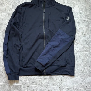 Svart vindjacka från CIRKL Workwear XL - Snygg svart vindjacka från CIRKL Workwear i storlek XL. Jackan har hel dragkedja, hög krage och coola mörkblå detaljer på ärmarna. Tillverkad i polyester, perfekt för dig som gillar stilren och sportig look. CIRKL-logga på ena ärmen för extra edge.