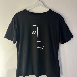 Svart t-shirt med minimalistiskt ansikte - Svart t-shirt i bomull och polyester med ett vitt minimalistiskt ansikte tryckt på framsidan. Klassisk rund hals och korta ärmar. Perfekt för dig som gillar stilren och konstnärlig design.