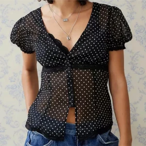 Svart prickig blus från Urban outfitters - Supersöt svart transparent blus med vita prickar, v-ringning och spetsdetaljer. Korta puffärmar med resår och knäppning framtill. Perfekt att styla med jeans eller kjol för en trendig look.