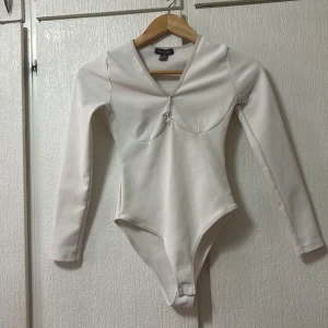 Vit långärmad bodysuit från Primark - Säljer en stilren vit långärmad bodysuit från Primark. Den har en v-ringad design med en liten knutdetalj framtill. Perfekt för att skapa en elegant look. Tillverkad i Turkiet.