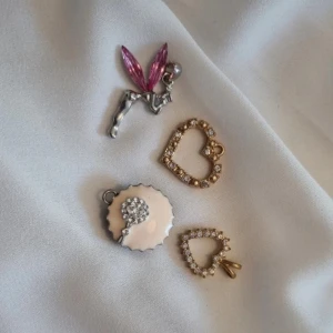 Charmiga berlocker i olika former - Säljer ett set med fyra unika berlocker. En silverfärgad älva med rosa vingar, två hjärtformade berlocker i guld med glittrande stenar, och en cupcakeformad berlock med en vit blomma. Perfekta för att ge dina smycken en personlig touch! De kostar 10 kr styck