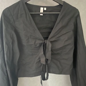 Svart knytblus - Snygg svart blus med knytning framtill och v-ringad design från Nelly. Blusen har långa ärmar och en croppad passform. 🖤🖤