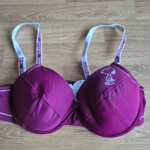 Helt ny bh med Snoopy-design 80A - Snygg vinröd bh med en söt Snoopy-design på kupan. De delvis glittriga axelbanden är justerbara och har Snoopy-text på sig. Uttagbara inlägg. Aldrig använd, prislapp kvar. Kommer från ett konkurslager. Se även mina andra auktioner för möjlighet till bundle med 20% rabatt.