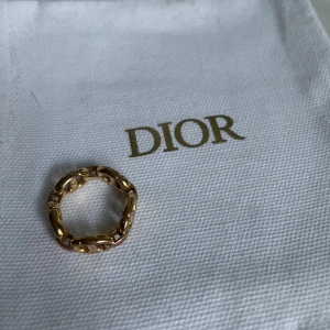 Guldfärgad ring från Dior - Elegant guldfärgad ring från Dior med en unik kedjedesign. Perfekt för att ge en lyxig touch till din stil. Ringen har en subtil märkning på insidan som visar att den är äkta.