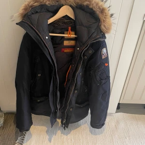 Svart vinterjacka från Parajumpers - Säljer en svart vinterjacka från Parajumpers med pälsfodrad huva och flera praktiska fickor. Jackan har en broderad patch på ärmen och dragkedja framtill. Perfekt för kalla dagar!