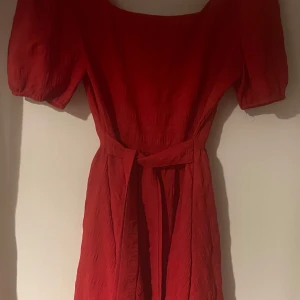 Röd klänning med puffärmar och öppen rygg - Snygg röd klänning med puffiga kortärmar och knytband i midjan. Perfekt för en somrig dag eller en festlig kväll. Klänningen har en elegant och feminin stil.