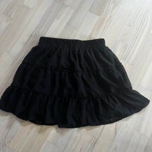 Svart volangkjol - Säljer en söt svart kortkjol med volanger. Kjolen har en elastisk midja för en bekväm passform. Perfekt för att styla med olika toppar och accessoarer.