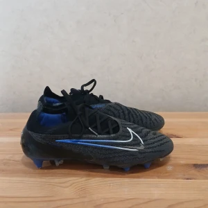 Nike phantoms Elite SG - Snygga svarta fotbollsskor från Nike med blå och vita detaljer. Skorna har snörning och en mönstrad yta för bättre grepp och kontroll. Perfekta för fotbollsplanen med sina robusta dobbar. Är i Elite prisklassen och medför extra kvalitet och metaldobbar. Storlek 40.5. Nypris över 2500.