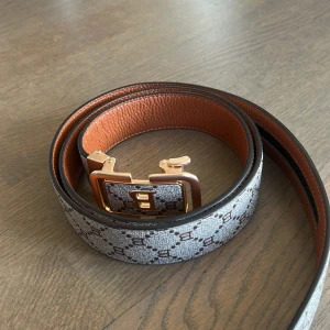 Mönstrat bälte med gulddetaljer - Snyggt bälte med ett klassiskt mönster i beige och brunt. Bältet har en iögonfallande guldfärgad spänne med bokstaven 'B'. Insidan är i en kontrasterande brun färg.