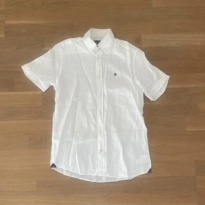 Vit linneskjorta från Morris - Säljer en stilren vit kortärmad linneskjorta från Morris. Skjortan har en broderad detalj på bröstet och knappar framtill. Perfekt för varma dagar med sin lätta och luftiga känsla.