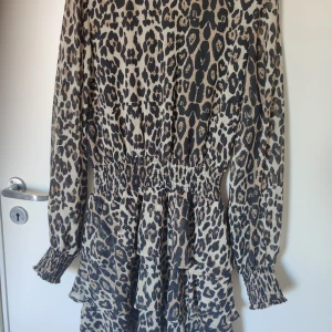 Leopardmönstrad klänning med volanger - Snygg leopardmönstrad klänning med volanger och långa ärmar. Perfekt för en trendig look. Den har en smockdetalj i midjan som ger en fin passform.