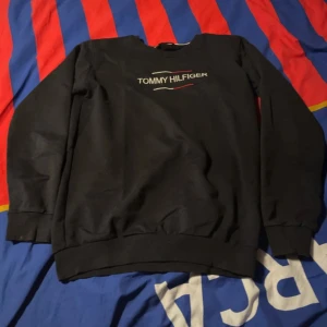 Svart sweatshirt från Tommy Hilfiger - Säljer en svart sweatshirt från Tommy Hilfiger med deras logga på bröstet. Tröjan har långa ärmar och är tillverkad i bomull för en bekväm passform. Perfekt för en stilren och avslappnad look. Oanvänd. Säljer pga att jag aldrig har använt den.