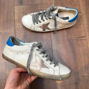 Golden goose skor - Snygga vita sneakers från Golden Goose med en distinkt stjärndetalj i brunt på sidan. Skorna har en blå häl och grå snörning med märkets logga. Sulan har en vintage-look med en lätt sliten finish.