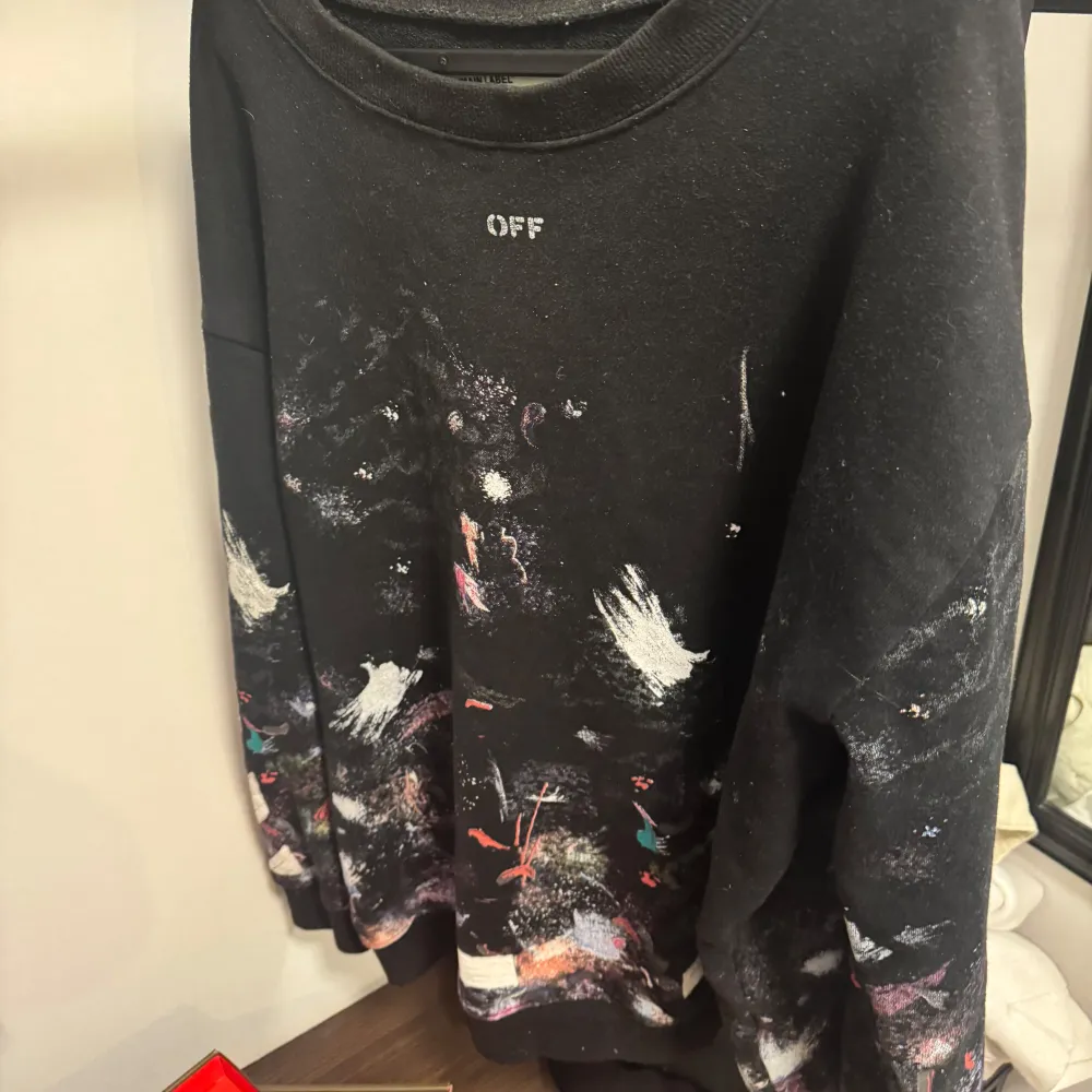Säljer en svart tröja från Off-White med unikt färgstänk och tryckdetaljer. Tröjan har långa ärmar och ett stort vitt tryck på baksidan. Perfekt för dig som gillar streetwear och vill sticka ut.. Neuletakit & Villapaidat.
