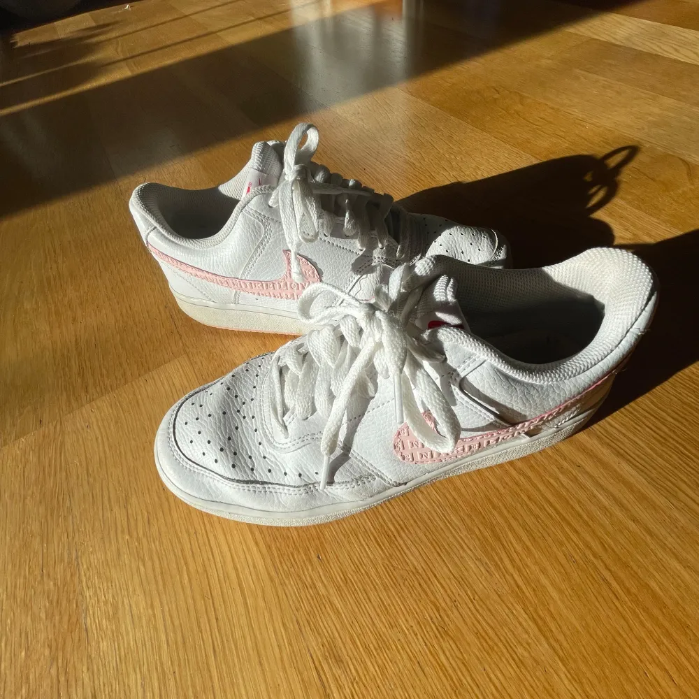 Säljer ett par vita Nike sneakers med rosa detaljer och snörning. Skorna har en klassisk design med perforeringar på tån för ventilation. Perfekta för en avslappnad stil.. Kengät.