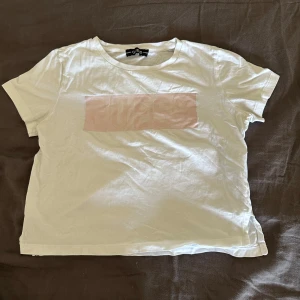 Vit t-shirt från Guess -  en vit t-shirt från Guess med ett subtilt rosa tryck på framsidan. T-shirten är kortärmad och tillverkad i bomull, vilket gör den bekväm och lätt att bära. Perfekt för en avslappnad stil. 