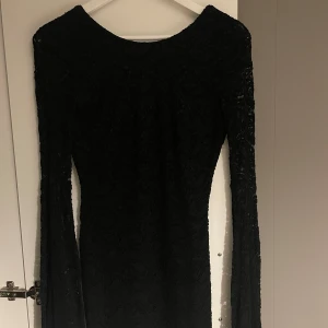 Svart långärmad klänning med spetsdetaljer - Figurnära elegant svart klänning med lång ärm och vacker spetsdesign. Klänningen har öppen rygg. 