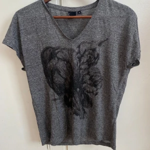 Grå t-shirt med tryck - Säljer en grå t-shirt med ett konstnärligt tryck på framsidan. T-shirten har en v-ringad hals och korta ärmar. Perfekt för en avslappnad stil.