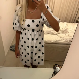 Vit klänning med svarta prickar - Charmig vit klänning med stora svarta prickar och puffiga kortärmar. Den har en fyrkantig urringning och en luftig passform som ger en lekfull look. Perfekt för sommarens alla tillfällen!