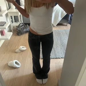 Svarta bootcut jeans med detaljer - Snygga svarta bootcut jeans med dekorativa detaljer vid fickorna. Perfekta för en stilren look. De har en låg midja och en klassisk bootcut-stil som ger en tidlös känsla. Endast använda 1 gång, de är i fint skick ❤️färgen syns bäst på andra bilden! Skriv vid funderingar💕i storleken 34 tall, lägg prisförslag!