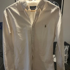 Vit skjorta från Polo Ralph Lauren - Klassisk vit skjorta från Polo Ralph Lauren med broderad logotyp på bröstet. Skjortan har långa ärmar och knappar framtill. Perfekt för en stilren look.