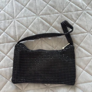 Svart glittrig handväska - Snygg svart handväska med glittrande detaljer och dragkedja. Perfekt för att ge en extra touch till din outfit. Väskan har ett bekvämt handtag och är lätt att bära med sig.