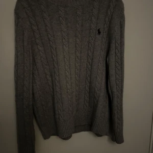 Grå kabelstickad tröja från Ralph Lauren - Säljer en stilren grå kabelstickad tröja från Ralph Lauren. Tröjan har en klassisk design med rund hals och långa ärmar. Perfekt för kyligare dagar och ger en tidlös look. 🐴