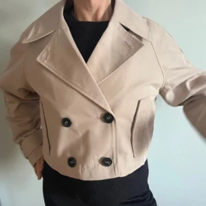 Beige trench med knappar - Snygg beige kappa med dubbelknäppning och stora svarta knappar. Jackan har en klassisk krage och är croppad i modellen. Perfekt för en stilren look.