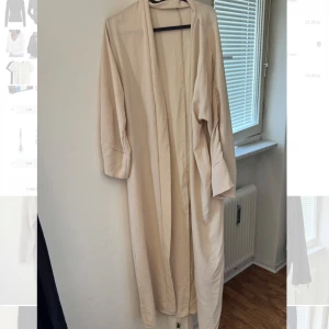 Beige lång trenchcoat / abaya - Säljer en elegant beige lång kappa med normal passform. Går även att använda som en abaya. Den har långa ärmar och en stilren design utan knappar eller dragkedja. Perfekt för en stilren look.