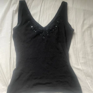 Svart topp från H&M - Snygg svart ärmlös topp från H&M med v-ringning och dekorativa paljetter vid halsen. Perfekt för en stilren look. Tillverkad i ett mjukt material som känns skönt mot huden.
