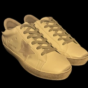 Beige sneakers - Snygga beige sneakers med en stjärndetalj på sidan. Skorna har snörning och en kontrasterande svart hälflik. Perfekta för en stilren look. Säljer för att dom var för småa för mig. Dock en replika! Där av billigt pris.