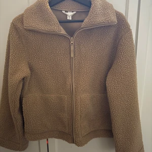 Beige teddyjacka med dragkedja - Mysig beige teddyjacka med dragkedja framtill. Perfekt för kyliga dagar med sin fluffiga och varma design. Jackan har en hög krage och långa ärmar för extra värme.