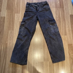 Grå cargobyxor med fickor - Säljer ett par grå cargobyxor med praktiska fickor på sidorna. Byxorna har en dragkedja och knapp framtill. Perfekta för en avslappnad stil. Jeansen är ifrån H&M.