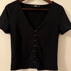 Svart kortärmad topp från BikBok - Snygg svart topp från BikBok med korta ärmar och v-ringning. Toppen har knappar framtill och är perfekt för en stilren look. Passar bra till både jeans och kjol.