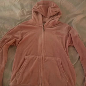 Rosa hoodie från Juicy Couture - Mysig rosa hoodie från Juicy Couture i färgen Pink Nectar. Tillverkad i mjuk polyester med dragkedja framtill och huva. Perfekt för en avslappnad stil.