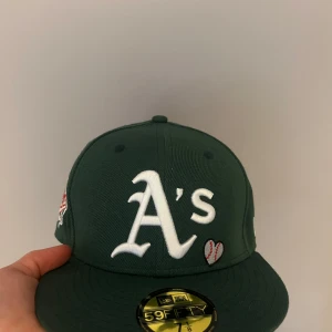 Grön keps från New Era (56,8cm) - Snygg grön keps från New Era med broderad logotyp och textdetaljer. Storlek är 56,8cm (7 1/8). Pris kan diskuteras.