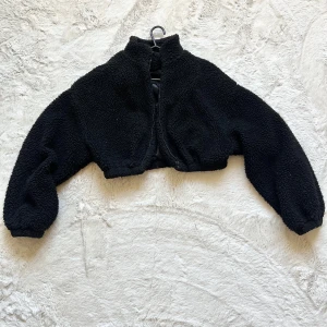 Svart teddyjacka - Säljer en mysig svart teddyjacka med croppad passform. Jackan har långa ärmar och en hög krage. Perfekt för kyliga dagar när du vill vara både varm och stilren.