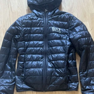 Svart pufferjacka från EA7 - Snygg svart pufferjacka från EA7 med huva och dragkedja framtill. Jackan har ett quiltat mönster och är perfekt för kyligare dagar. Den har långa ärmar och en stilren design med logotyp på bröstet.
