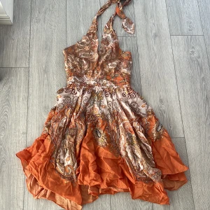 Orange mönstrad klänning - Säljer en färgglad klänning i orange med ett vackert paisleymönster. Klänningen har en omlott-design och knytband vid halsen. Perfekt för sommarens festligheter! 🌸insydd i urringning men kan sprättas upp om man önskar 