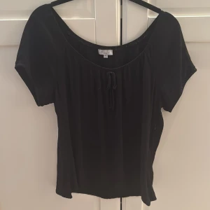 Svart kortärmad topp från Ell Woman - Säljer en stilren svart topp från Ell Woman. Den har korta ärmar och en söt knytdetalj framtill. Perfekt för en enkel och elegant look. I storlek 42