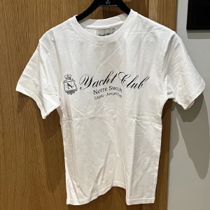 Vit t-shirt med tryck - Snygg vit t-shirt med texten 'Yacht Club Notre Succès' tryckt på framsidan. Perfekt för en avslappnad stil. Kortärmad och tillverkad i mjukt material för extra komfort.