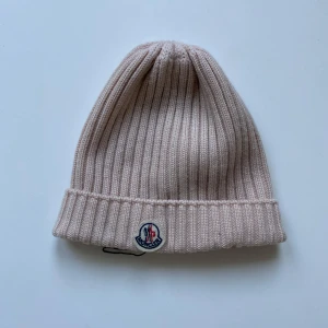 Moncler mössa - Säljer en snygg beige ribbad mössa från Moncler i storlek M. Perfekt för kyliga dagar med sin stilrena design och varma material. Mössan har Monclers ikoniska logga framtill. Passar perfekt till både höst och vinteroutfits!