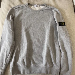 Grå sweatshirt från Stone Island - Säljer en stilren grå sweatshirt från Stone Island. Tröjan har en klassisk rund hals och långa ärmar. Den är dekorerad med det ikoniska Stone Island-märket på ärmen. Perfekt för en avslappnad och trendig look.