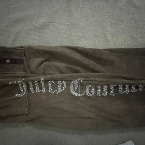 Bruna byxor från Juicy Couture - Snygga bruna byxor från Juicy Couture med glittrande text längs benet. Byxorna har en loose passform och är tillverkade i ett mjukt material som ger en bekväm känsla. Perfekta för en avslappnad stil med en touch av glamour.