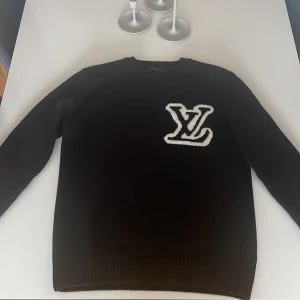 Svart tröja med logotyp - Snygg svart tröja från Louis Vuitton med en stor vit logotyp på bröstet. Tröjan har långa ärmar och ribbade kanter vid ärmslut och nederkant. Perfekt för en stilren look.