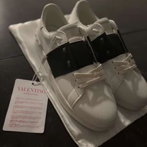 Valentino Garavani vita sneakers - Snygga vita sneakers från Valentino Garavani med en bred svart rem över ovansidan. Skorna har snörning och en stilren design med märkets logotyp på tungan och insidan. Perfekta för en trendig look. Skorna är sprillans nya och passade tyvärr inte efter jag skickat efter dem🙁påse ingår samt kvitto, storlek 45